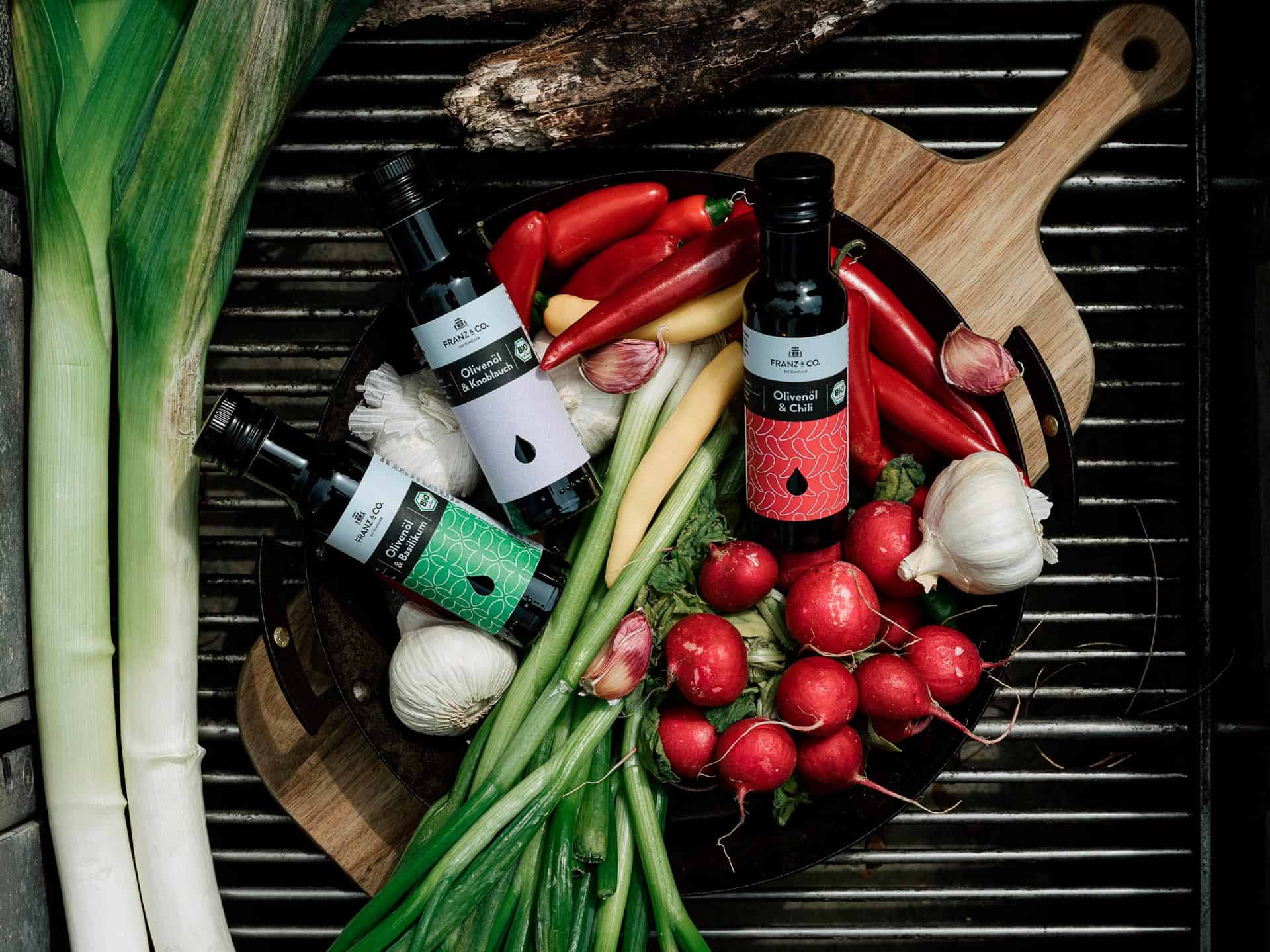 Olivenöl mit Chili, Knoblauch oder Basilikum von FRANZ & CO. liegen in einer Schale mit frischem Gemüse bereit.